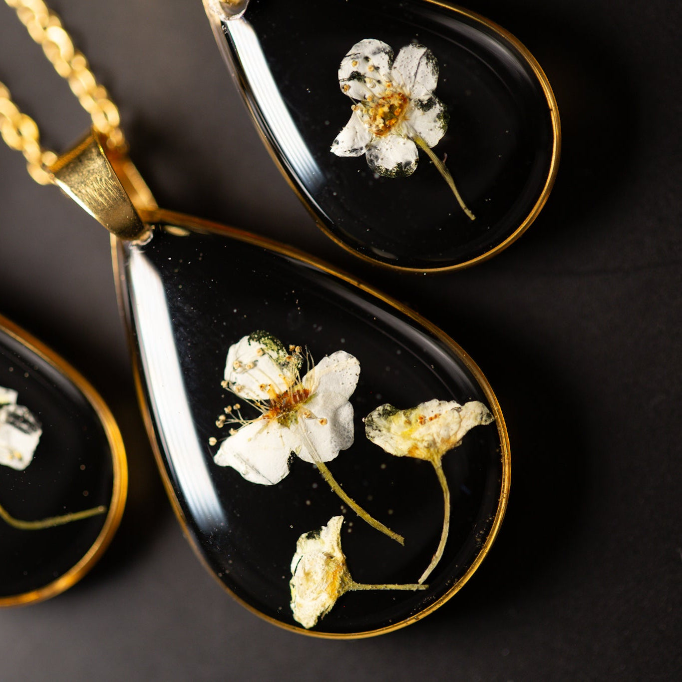Black teardrop pendants with gold accents and pressed flowers on a dark background