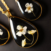 Black teardrop pendants with gold accents and pressed flowers on a dark background